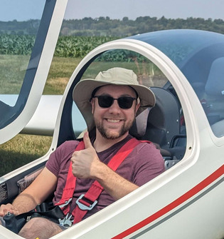 Andy sitting in a small two person glider airplane giving a thumbs up. The canopy is open and he has a hat on.